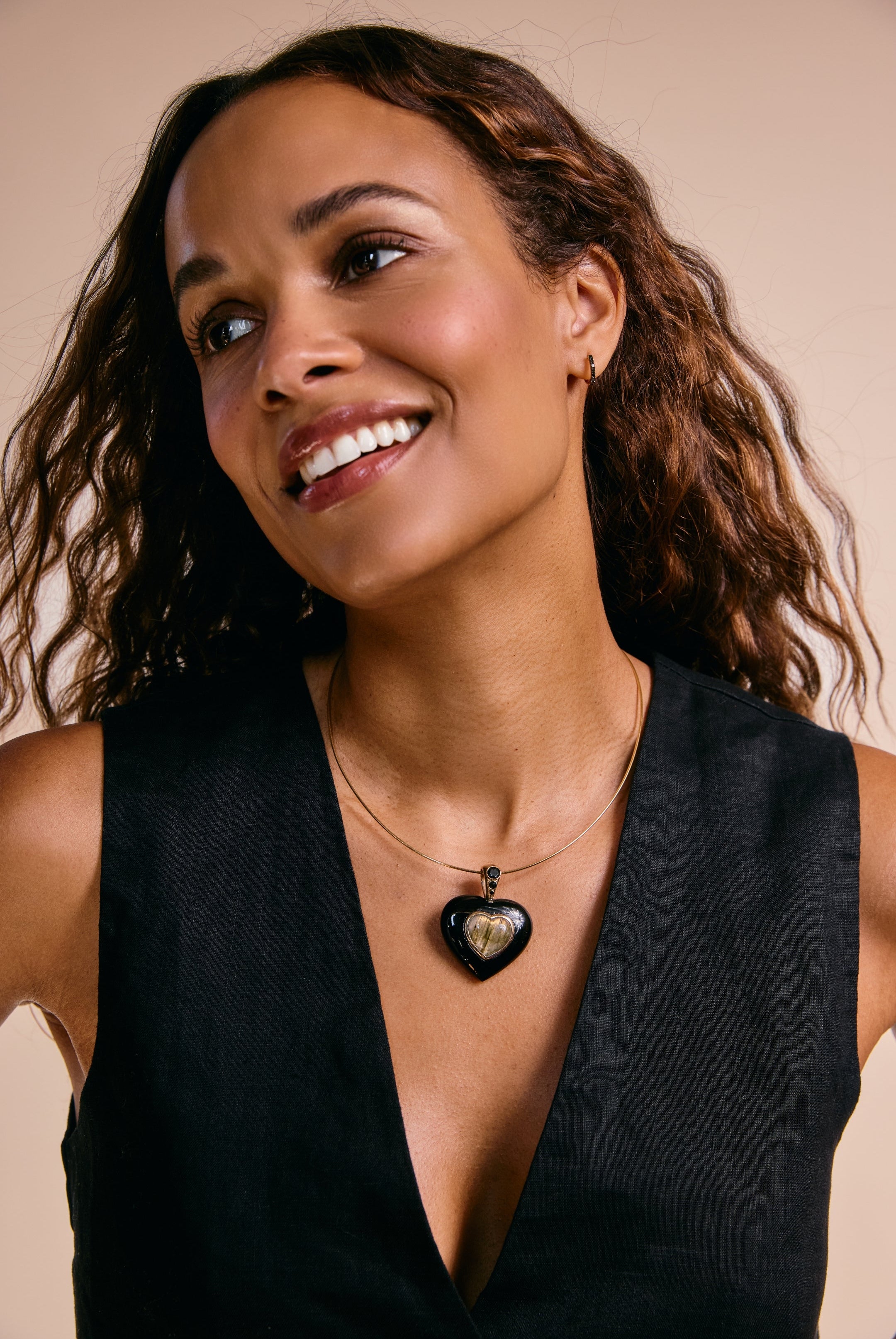 Woman wearing a black sleeveless top and a necklace with a heart pendant against a beige background